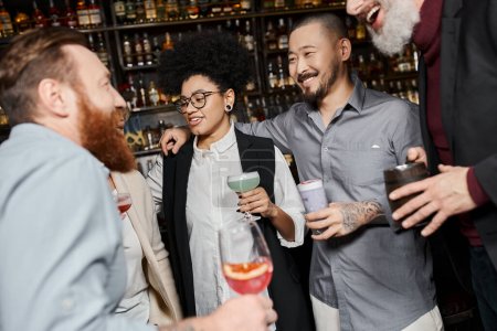 Téléchargez les photos : Compagnons de travail multiculturels tenant des lunettes et souriant pendant la conversation dans le bar à cocktails après le travail - en image libre de droit