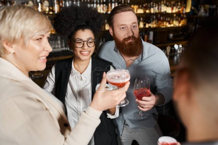 bärtiger tätowierter Mann und afrikanisch-amerikanische Frau lächeln in der Nähe von Arbeitskollegen mit Cocktailgläsern in Bar