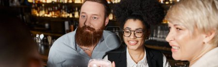 Téléchargez les photos : Homme barbu surpris près de femmes multiethniques gaies passer après le temps de travail dans un bar à cocktails - en image libre de droit