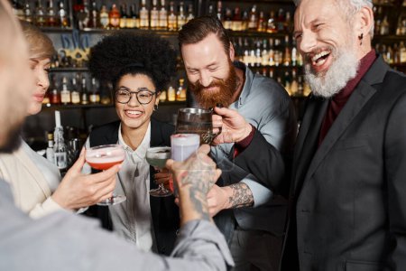 bearded tattooed men clinking glasses with happy multiethnic women in cocktail bar after work