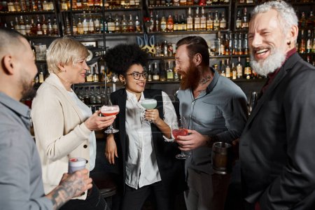 bärtiger tätowierter Mann spricht mit lächelnder afrikanisch-amerikanischer Frau in der Nähe von Arbeitskollegen bei Drinks in der Bar