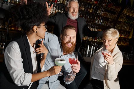 aufgeregter bärtiger Mann zeigt Siegeszeichen neben lachenden multiethnischen Freunden, die Cocktails in Bar halten