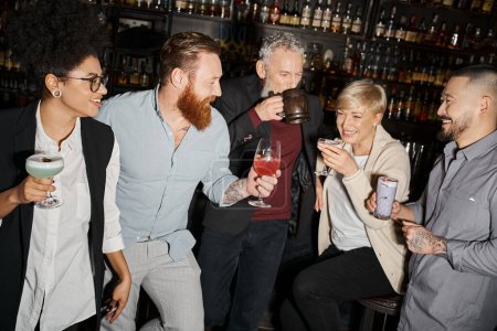 Téléchargez les photos : Homme tatoué barbu avec verre à cocktail près heureux compagnons de travail multiethnique dans le bar, amusant après le travail - en image libre de droit