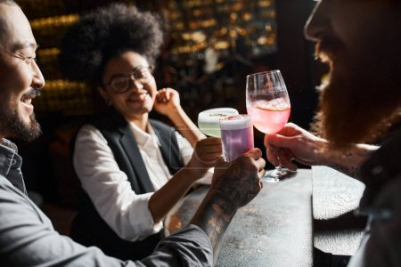 smiling african american woman clinking cocktail glasses with bearded tattooed colleagues in bar
