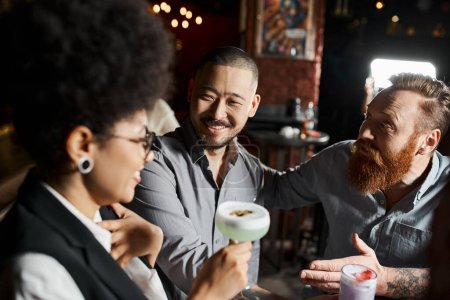 bärtiger tätowierter Mann im Gespräch mit überraschter afrikanisch-amerikanischer Frau neben asiatischem Kollegen in Bar