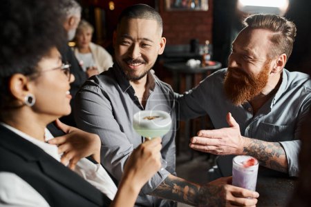 Téléchargez les photos : Barbu tatoué homme parler à étonné afro-américain femme près sourire asiatique collègue dans bar - en image libre de droit