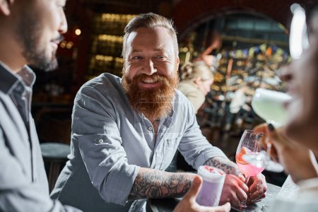 hombre barbudo tatuado con copa de cóctel sonriendo cerca de colegas multiétnicos en el bar, tiempo de fiesta