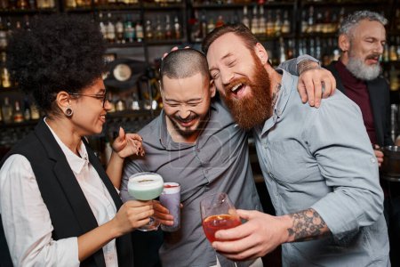 tätowierte multiethnische Kollegen mit Cocktails lachen neben fröhlicher afrikanisch-amerikanischer Frau in Bar