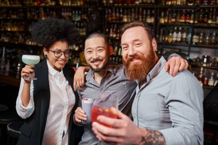 collègues multiethniques tenant des lunettes avec des cocktails et souriant à la caméra dans le bar, repos après le travail