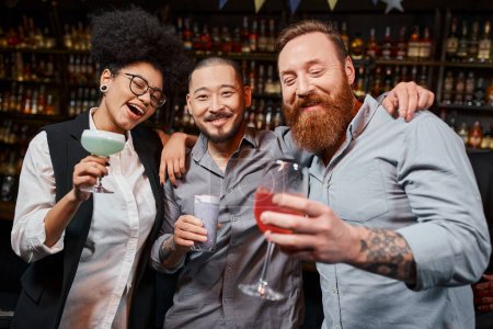 bärtiger asiatischer Mann blickt in die Kamera bei fröhlichen multiethnischen Freunden mit Cocktails in der Bar
