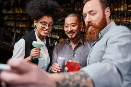 Photo for Tattooed bearded man taking photo on smartphone with happy multicultural workmates in cocktail bar - Royalty Free Image