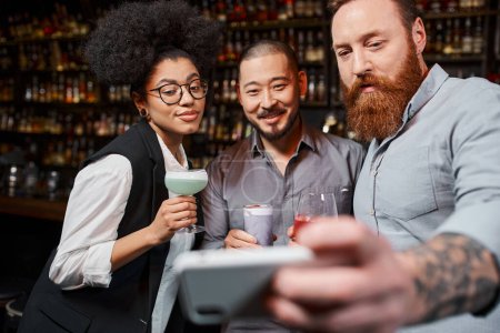 tattooed bearded man taking snapshot on smartphone with happy multiethnic work friends in bar