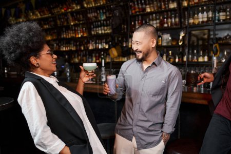 fröhliche asiatische Mann und afrikanisch-amerikanische Frau mit Drinks lächeln einander in Cocktailbar