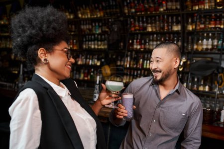 Africano americano mujer y asiático hombre con bebidas sonriendo durante conversación en cocktail bar,