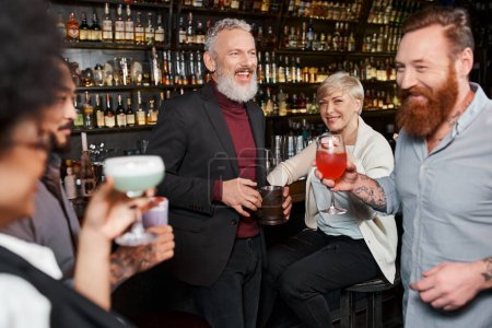 hombre barbudo feliz riendo cerca de colegas multiétnicos celebración de cócteles en el bar, tiempo de fiesta