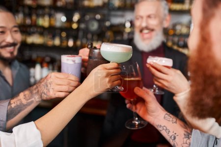 joyful multiethnic workmates clinking glasses with cocktails in bar after work, blurred background