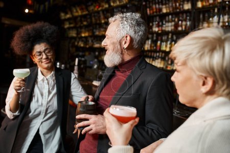 bärtiger Mann mittleren Alters lächelt neben multiethnischen Frauen mit Cocktails, Kollegen ruhen sich in Bar aus
