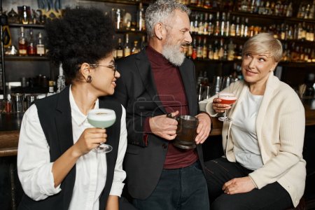 multiethnische Frauen mit Cocktailgläsern lächeln im Gespräch mit bärtigen Kollegen in der Bar