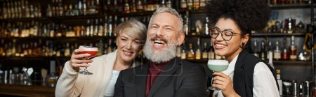 happy multiethnic women holding cocktail glasses near bearded colleague laughing in bar, banner