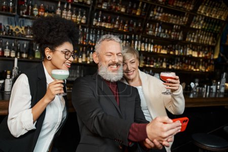 joyeuses femmes multiethniques avec des cocktails près de collègue barbu prendre selfie sur smartphone dans le bar