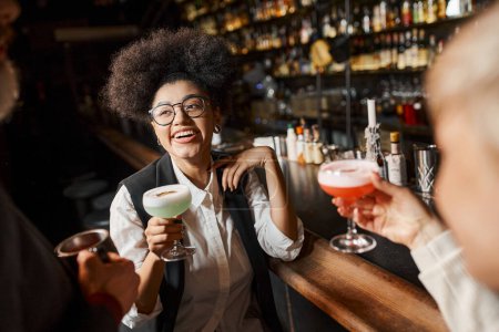 Téléchargez les photos : Femme afro-américaine gaie tenant verre à cocktail tout en passant du temps avec des collègues dans le bar - en image libre de droit