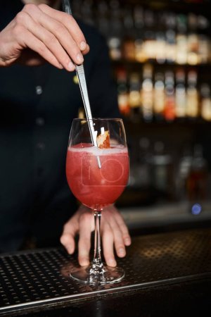 Téléchargez les photos : Vue recadrée du barman ajoutant une tranche d'orange fraîche dans un cocktail d'alcool de fruits sur le comptoir dans le bar - en image libre de droit
