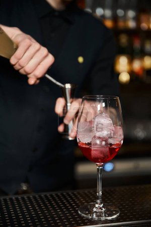 glass with alcoholic cocktail with ice cubes near cropped bartender working on blurred background
