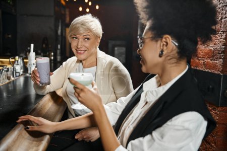smiling middle aged woman with african american colleague holding cocktails and talking in bar