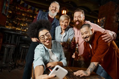 funny african american woman sticking out tongue and taking selfie with multiethnic workmates in bar