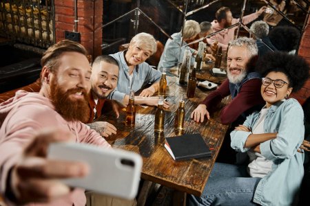 happy bearded man taking selfie with multiethnic colleagues near beer bottles on wooden table in pub