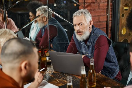 Téléchargez les photos : Homme barbu assis près d'un ordinateur portable et discuter du projet de démarrage avec l'équipe multiethnique dans le bar - en image libre de droit