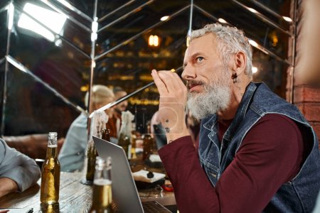 thoughtful bearded man looking away near laptop and colleagues reflecting in mirror in pub