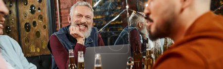 hombre barbudo feliz sentado junto a la computadora portátil y compañeros de trabajo multiétnicos hablando en el pub después del trabajo, pancarta