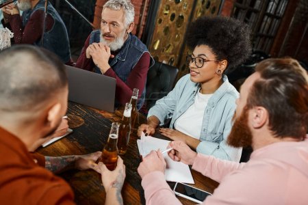Photo for Multiethnic business colleagues sitting in pub near laptop while planning new startup project - Royalty Free Image