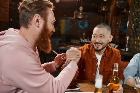joyful asian man shaking hands with bearded colleague while closing deal near beer bottles in pub