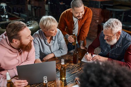 diverse multiethnic team looking at colleague writing in notebook during meeting in pub after work