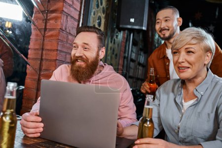fröhliches multiethnisches Team lächelt neben Laptop und Bierflaschen, während es nach Feierabend Zeit in der Kneipe verbringt
