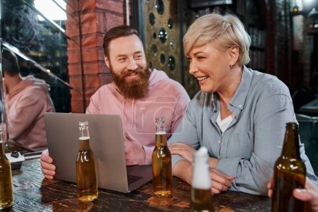 Lächelnder bärtiger Mann im Gespräch mit fröhlichen Kollegen mittleren Alters in der Nähe von Bierflaschen und Laptop in der Kneipe