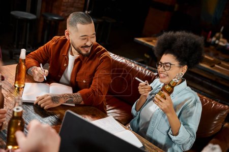 alegre africano americano mujer con cerveza botella cerca asiático colega sentado con notebook en pub