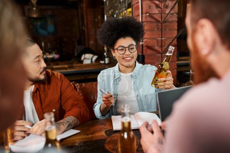 african american woman with beer bottle sharing idea of new startup with multiethnic team in  pub