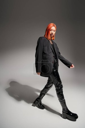 mujer asiática con estilo y confianza con el pelo rojo caminando en traje negro, pantalones de látex y chaqueta mirada
