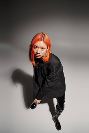 asian woman with red hair posing in black latex boots and blazer on grey background, high angle