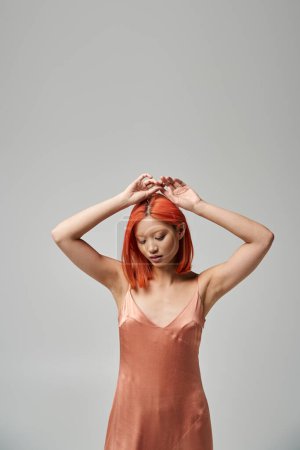 portrait of pretty young asian woman with red hair posing in silk slip dress on grey background