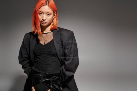 confident asian woman with red hair and piercing in nose posing in blazer on dark background