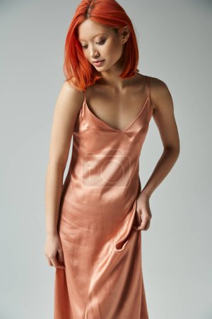 elegant young asian woman with red hair posing in silk and pastel pink slip dress on grey background