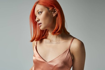 portrait of sensual young asian woman with red hair posing in slip dress on grey backdrop, feminine