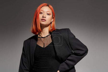 portrait of stylish asian and young woman in blazer looking at camera on grey background, eye makeup