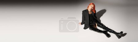 Téléchargez les photos : Pleine longueur de sassy asiatique femme en latex sexy bottes et blazer assis sur fond gris, bannière - en image libre de droit