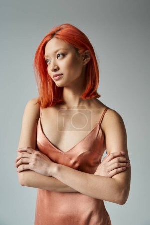 smiling young asian woman with red hair posing in pastel slip dress on grey backdrop, looking away