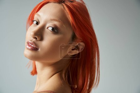 portrait of pretty asian woman with red hair looking at camera on grey background, feminine grace